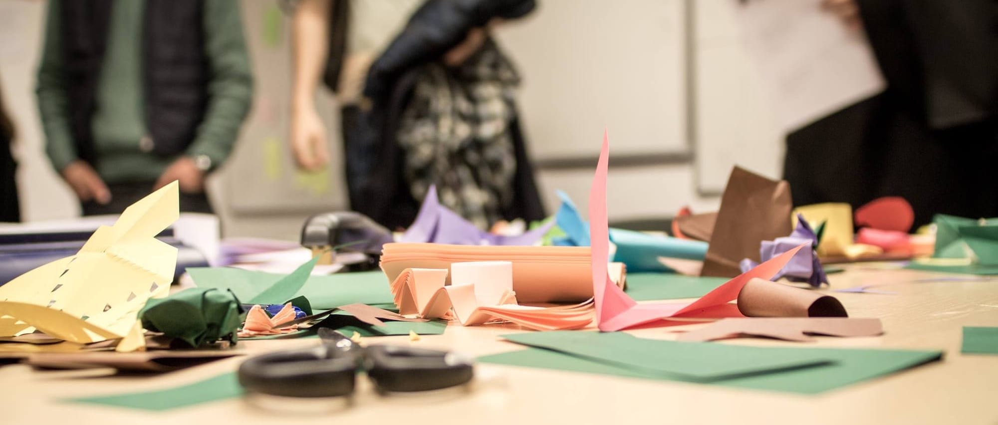 Image of a table with colourful papers, cardboards, scissor and staples. People is seen in the background, behind the table.